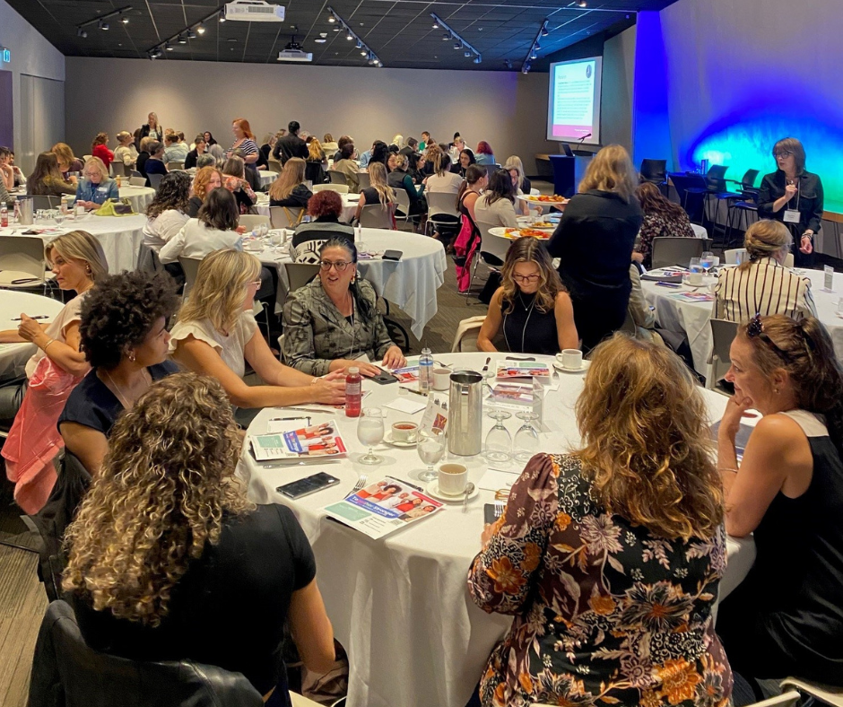 WeMB Business Breakfast attendees are seating at a large round table, smiling and chatting while other attendees mill about in the background.